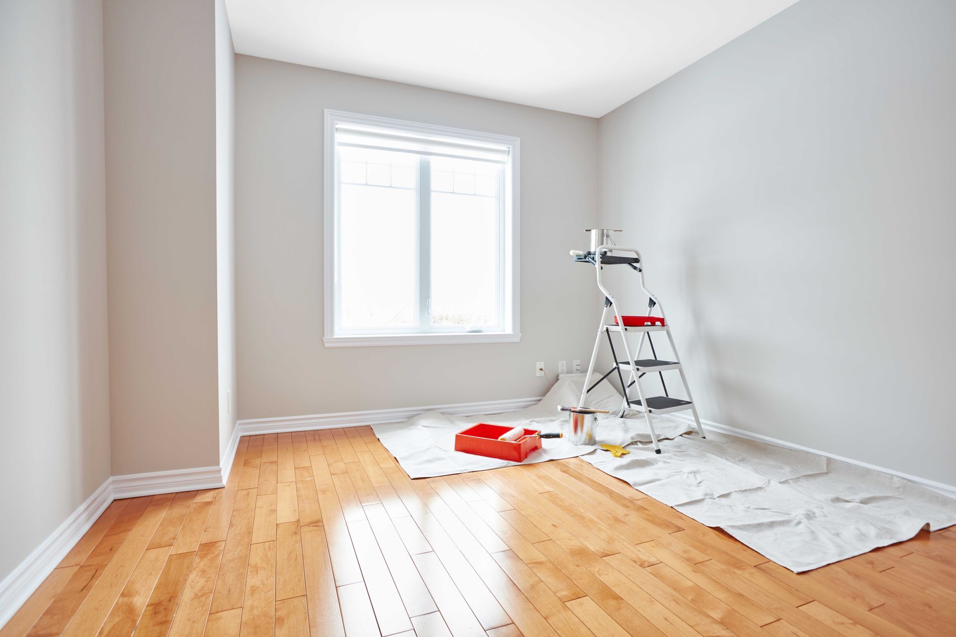 Chambre avec plancher de bois, murs gris clair fraîchement peints, fenêtre, échelle et fournitures de peinture.