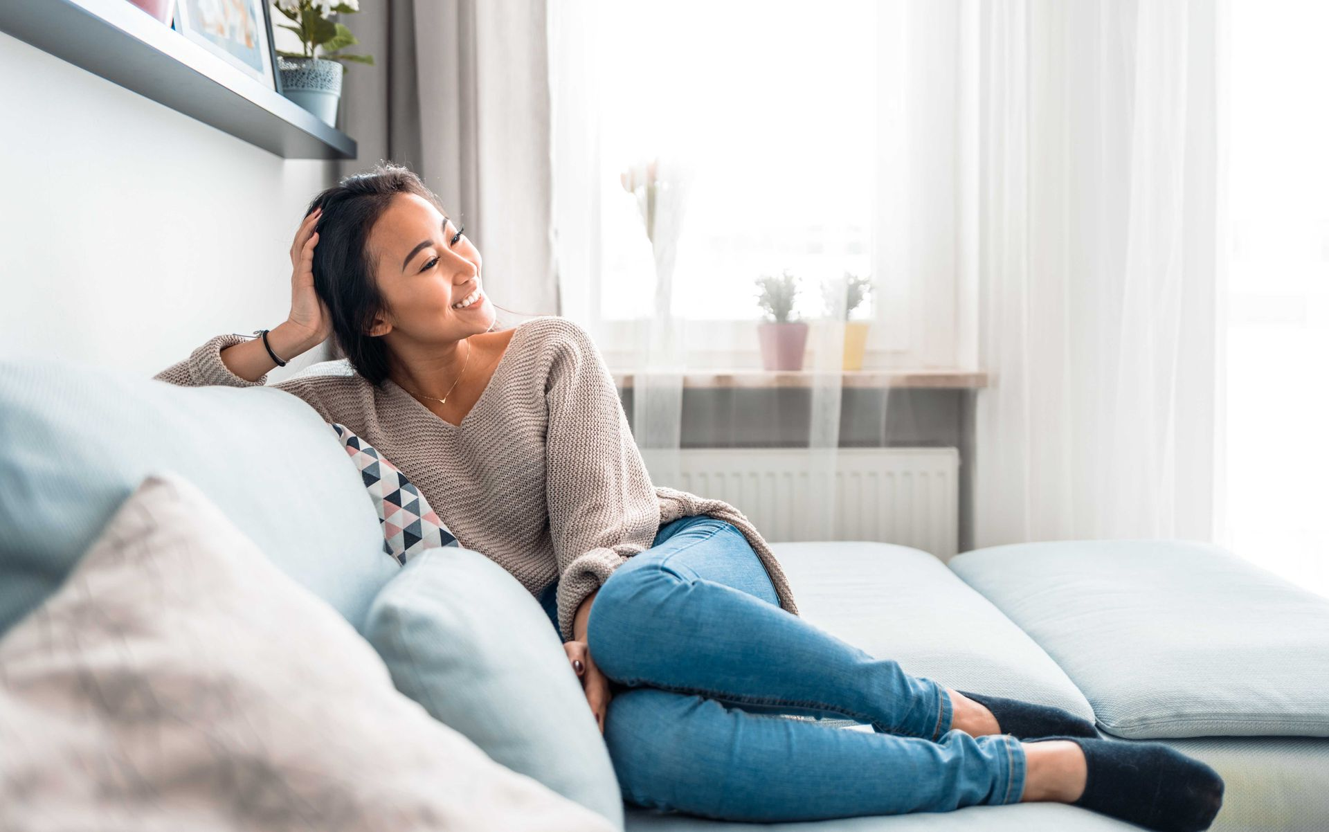 Une femme détendue sur un canapé bleu, souriante, regarde par la fenêtre. Elle porte un pull et un jean.