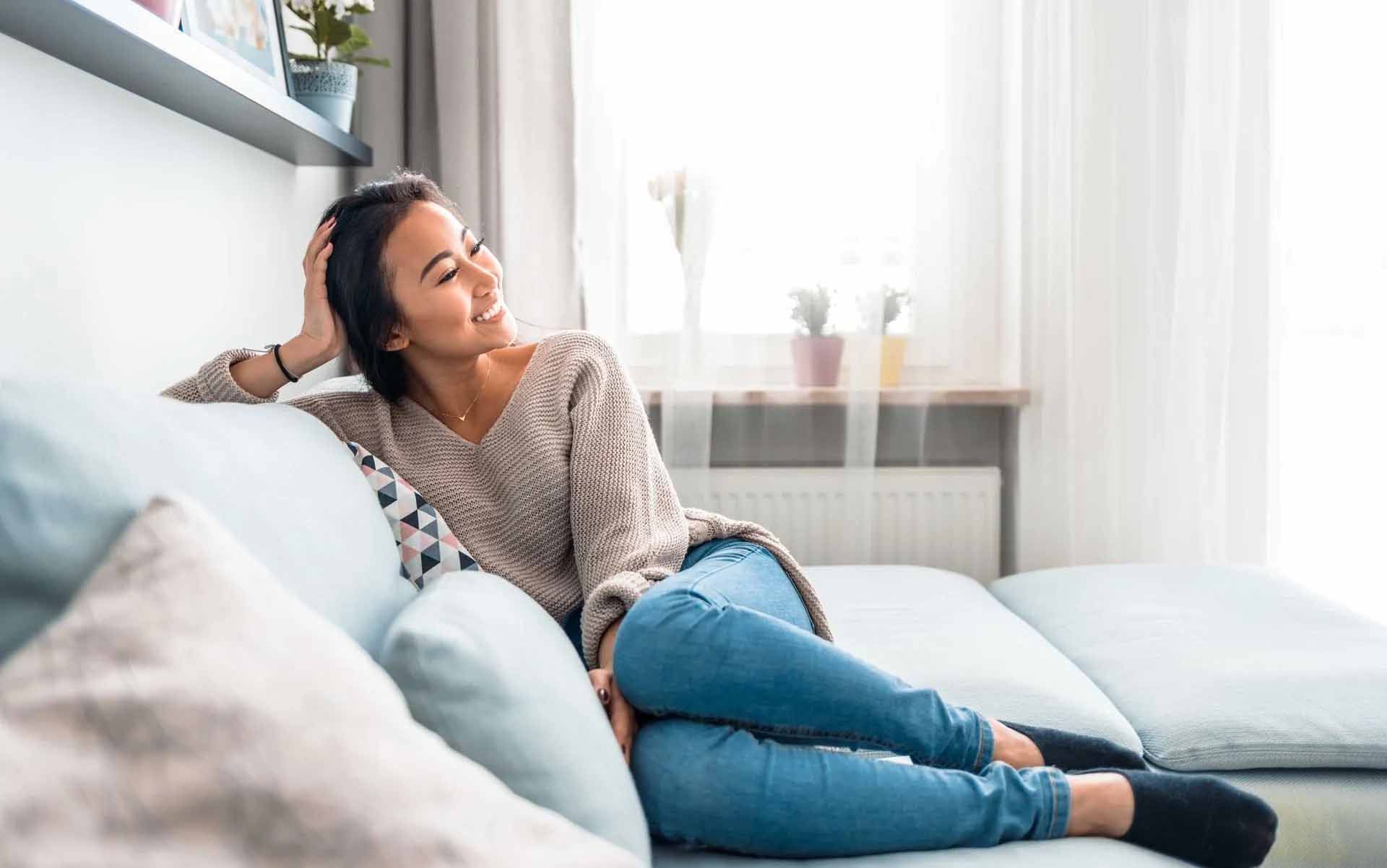 Une femme détendue sur un canapé bleu, souriante, regarde par la fenêtre. Elle porte un pull et un jean.