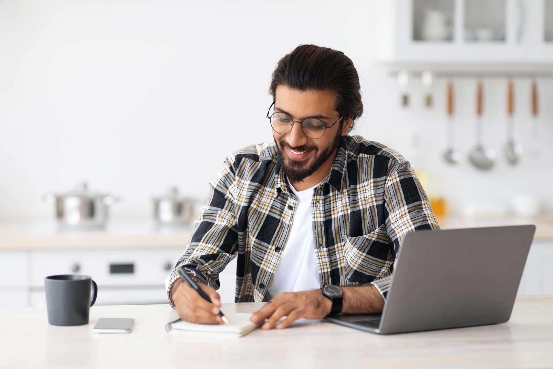 A man is sitting at a table with a laptop and writing in a notebook