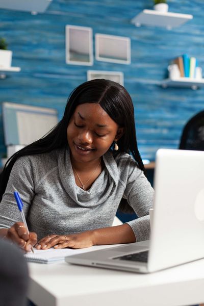 A woman is sitting at a desk with a laptop and writing on a piece of paper.