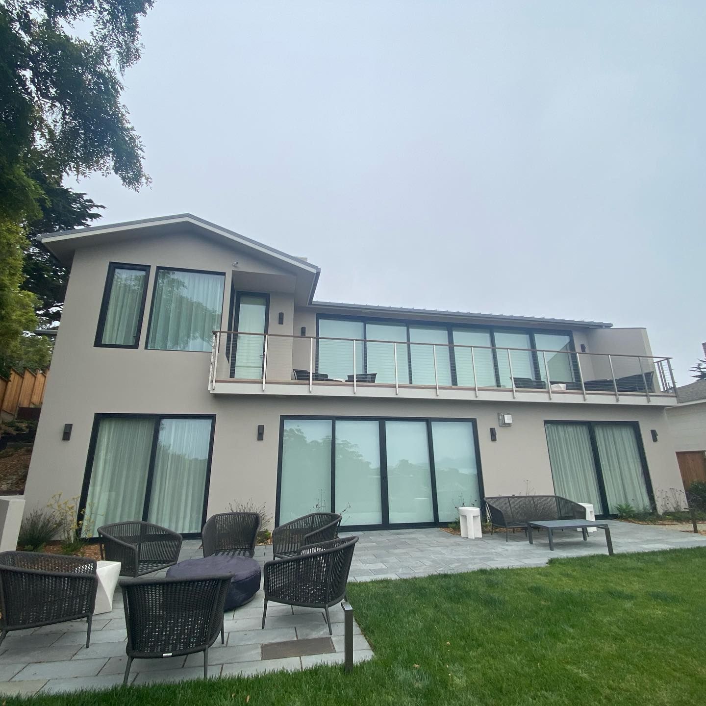 A light-colored two-story house with a glass balcony, floor-to-ceiling windows, and patio furniture on a stone terrace.