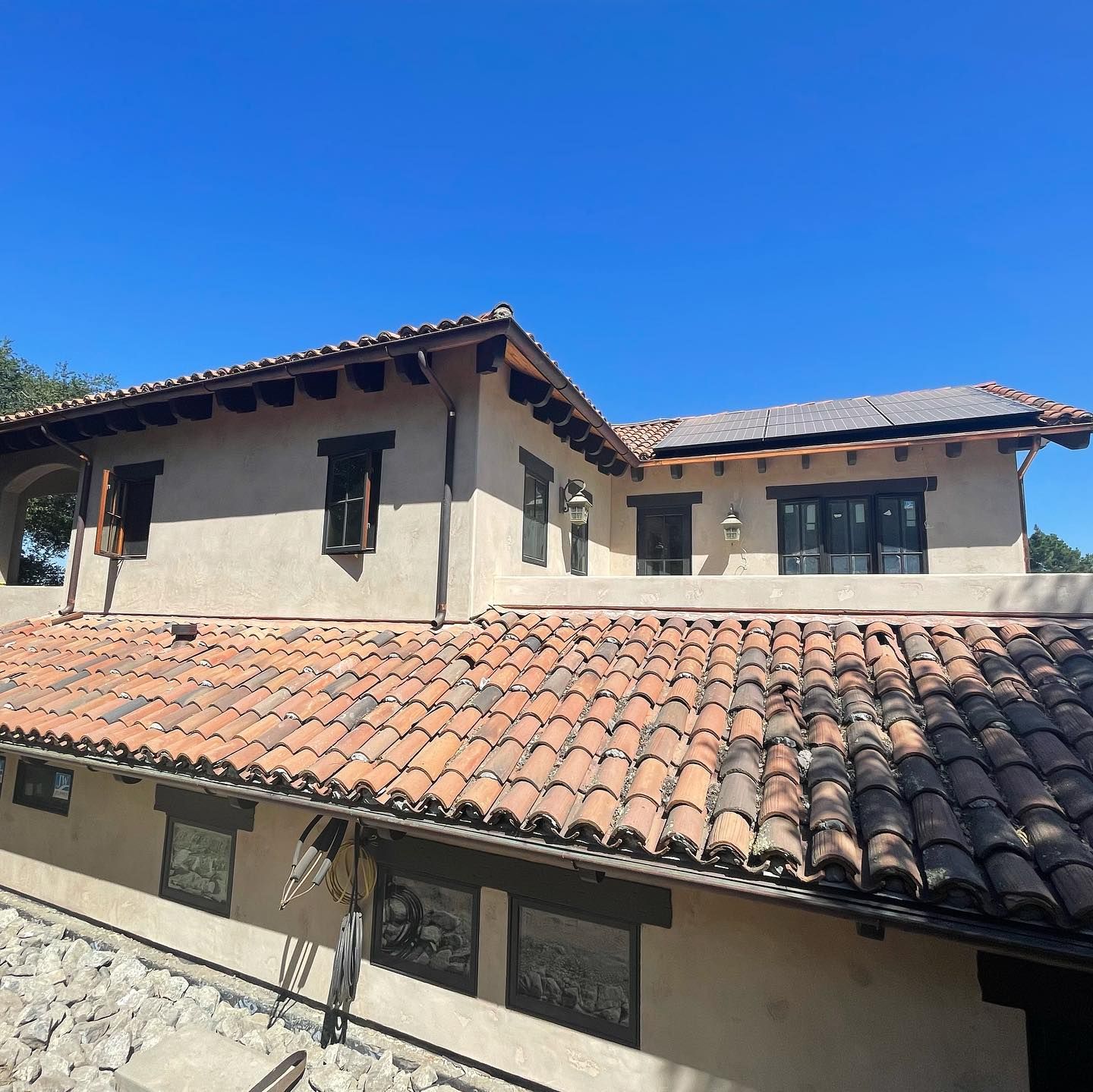 A two-story stucco home with a terracotta tile roof, wooden beams, and dark-framed windows under a clear blue sky.