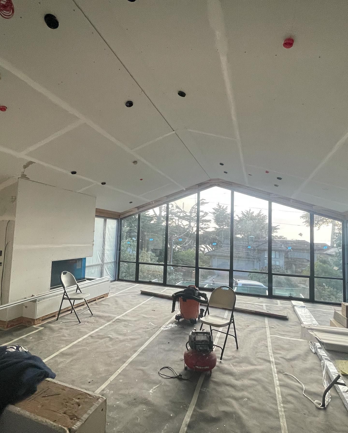 A room under construction featuring a vaulted ceiling, large wall-to-wall windows, and equipment on a covered floor.