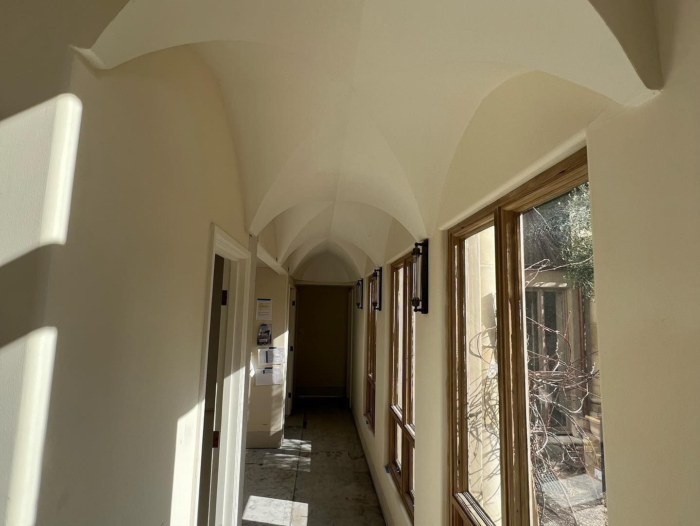 A sunlit indoor hallway with a white vaulted ceiling, light-colored walls, and large windows looking outside.
