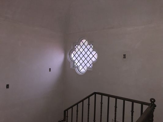 A clover-shaped window with a metal grid set in a plain, off-white wall above a dark metal staircase railing.