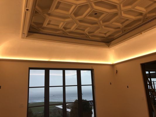 A room with a coffered ceiling, cove lighting, and a window overlooking a landscape and the sea.