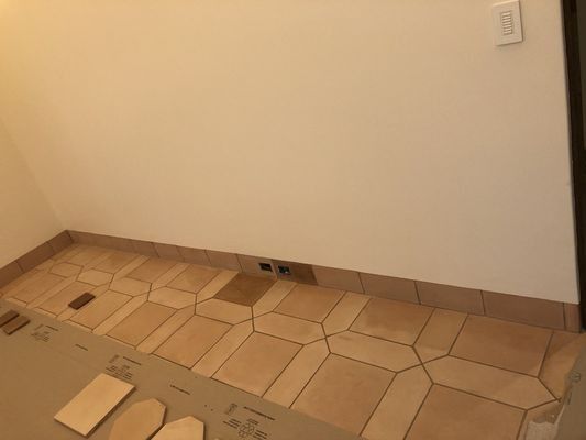 A partially tiled floor with light brown, geometric tiles along a white wall and a light switch on the upper right.