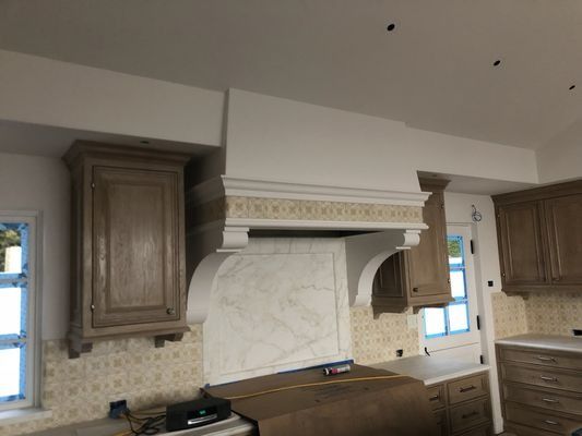A kitchen under construction featuring wooden cabinets, a large white decorative range hood, and marble backsplash tile.