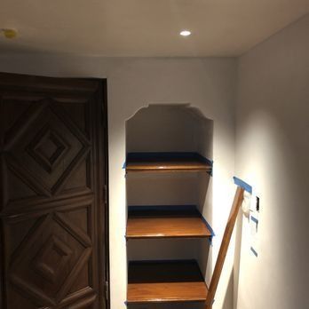 A recessed arched wall niche with three dark wooden shelves, flanked by a patterned wooden door and a wall with blue tape.