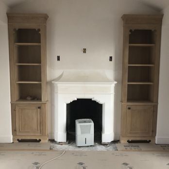 Two wooden floor-to-ceiling built-in cabinets flank a white fireplace mantel in an unfinished room with carpeted floors.