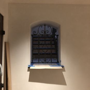 An arched window with a black metal decorative grate, showing a staircase in the background, set in a plain white wall.