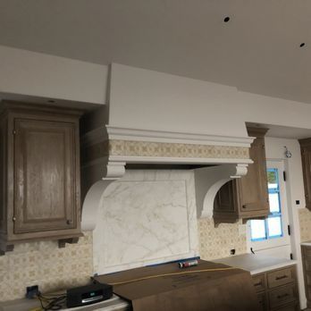 A kitchen under renovation features wood cabinets, a marble backsplash, and a prominent white range hood with tile detail.