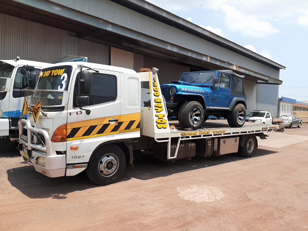 A white Hino tow truck carrying a blue Jeep Wrangler parked outside a building on a dirt surface. — Prompt Tilt Tray Towing & Recovery in Katherine, NT