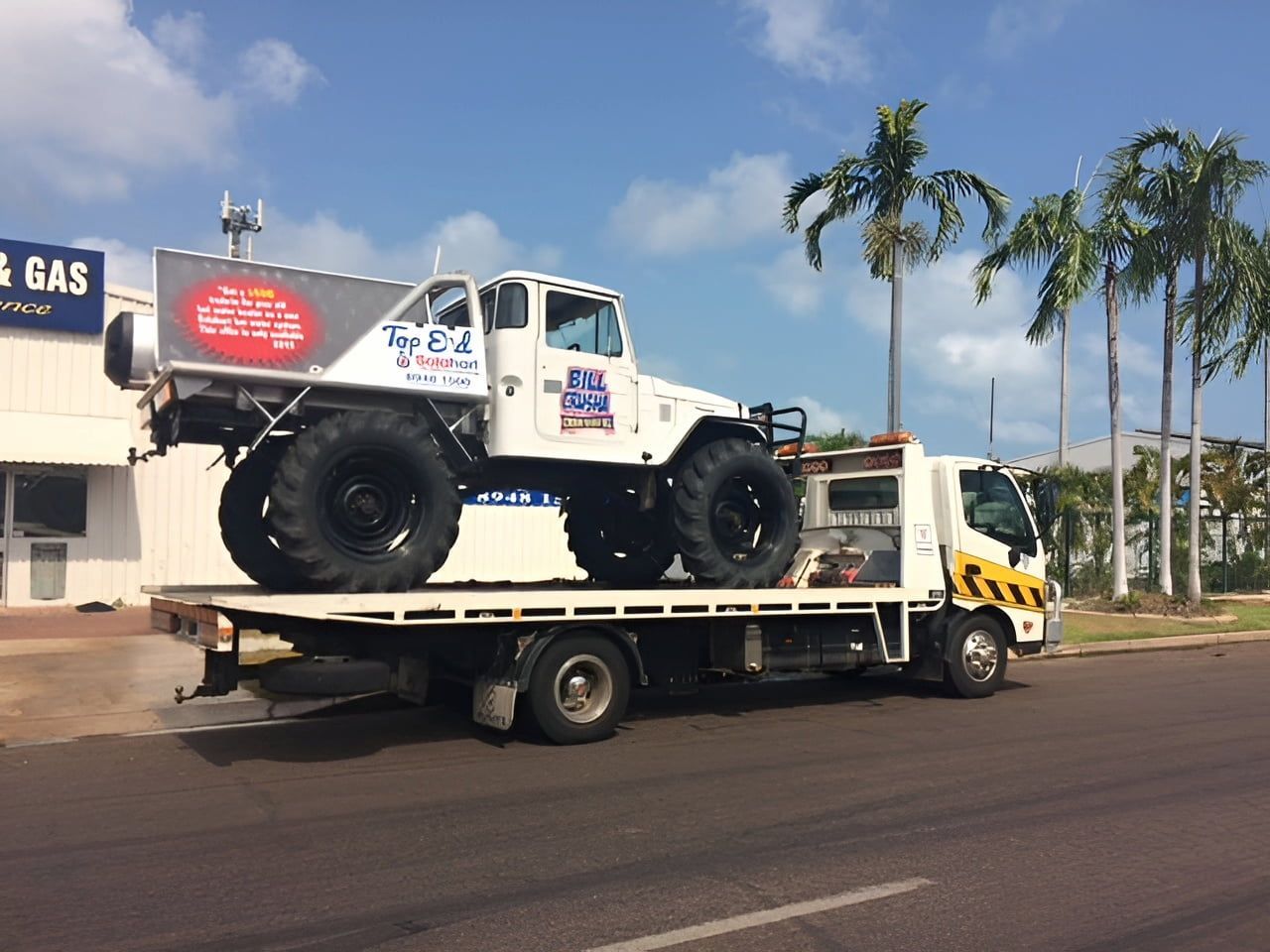 White off-road truck on a flatbed tow truck on a sunny street with palm trees — Prompt Tilt Tray Towing & Recovery in Herbert, NT