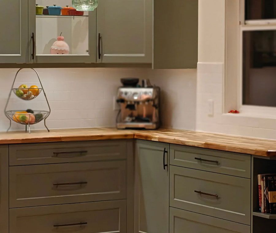 Green kitchen cabinets with wooden countertops, espresso machine, and fruit basket.