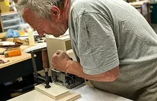 A person in a gray shirt works on wood in a workshop, using a clamp to hold pieces together.
