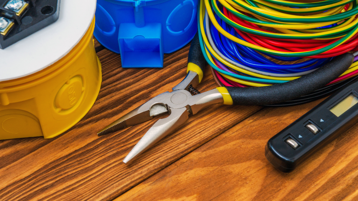 Electrical tools and wires on wood: pliers, multi-colored wires, a voltage tester, and electrical boxes.