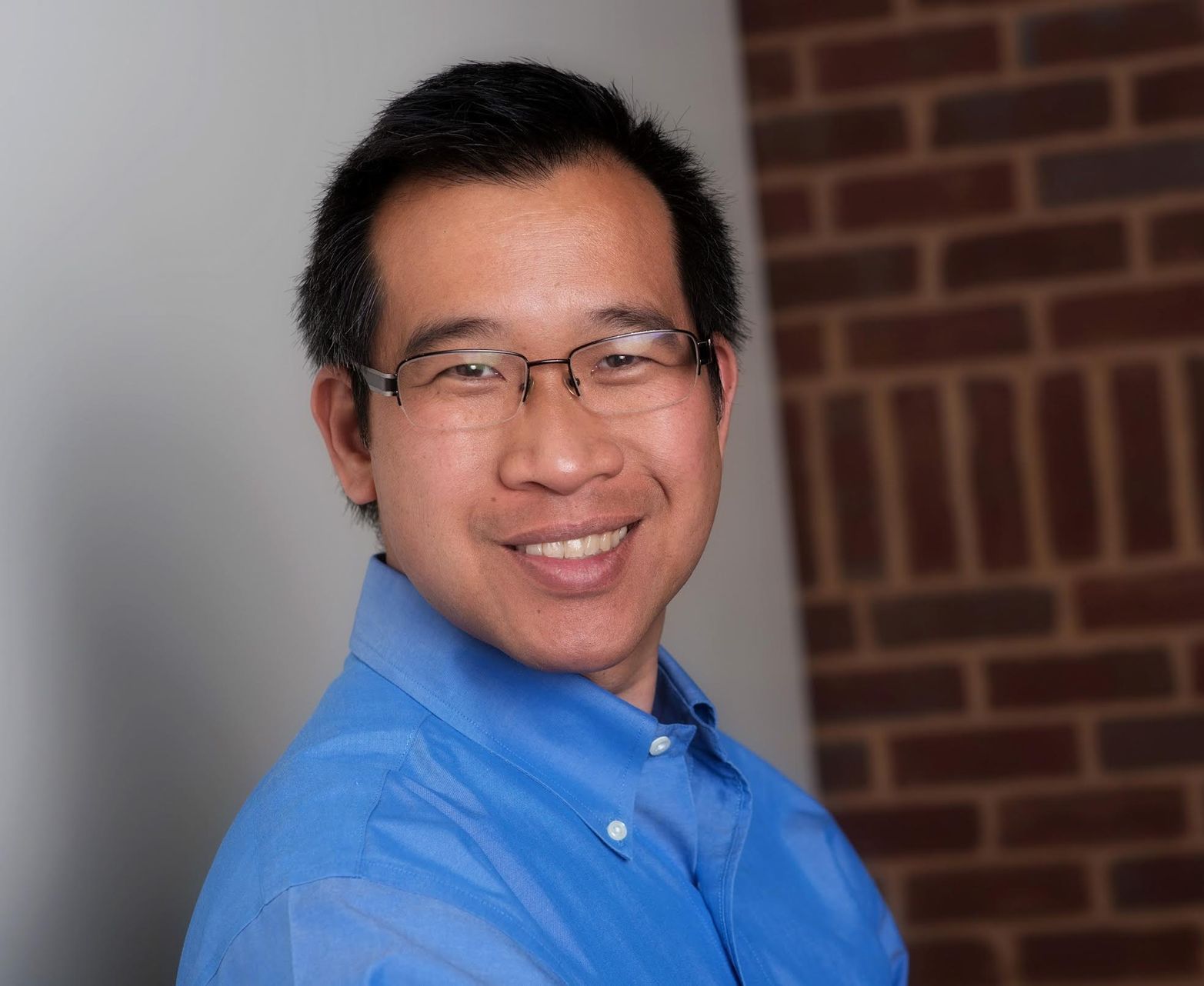 A man wearing glasses and a blue shirt is smiling in front of a brick wall.