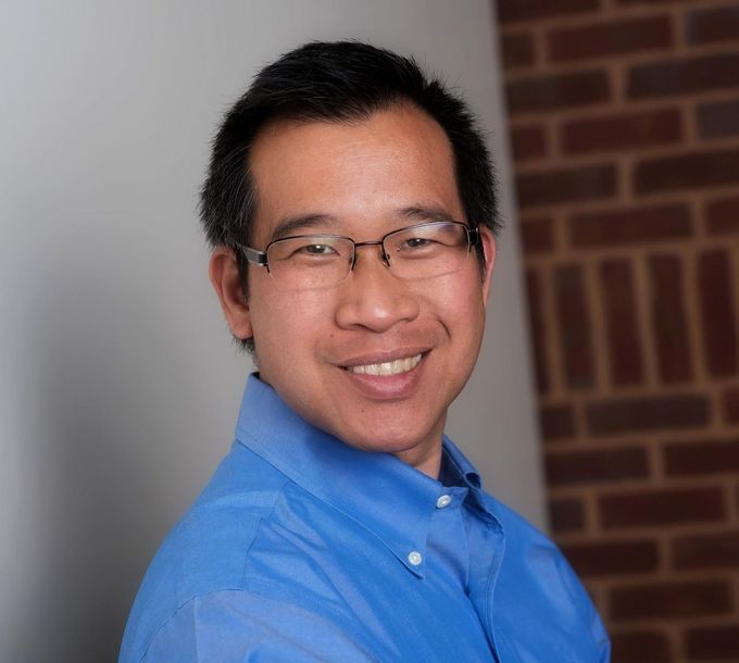 A man wearing glasses and a blue shirt is smiling in front of a brick wall.