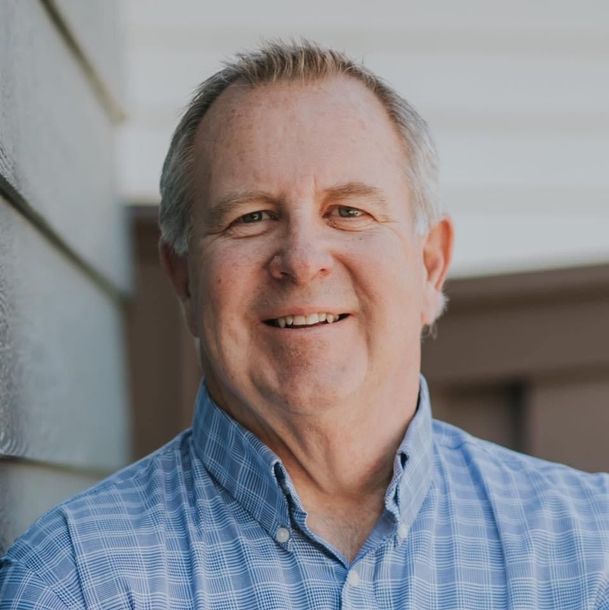 A man in a blue shirt is smiling for the camera while leaning against a wall.