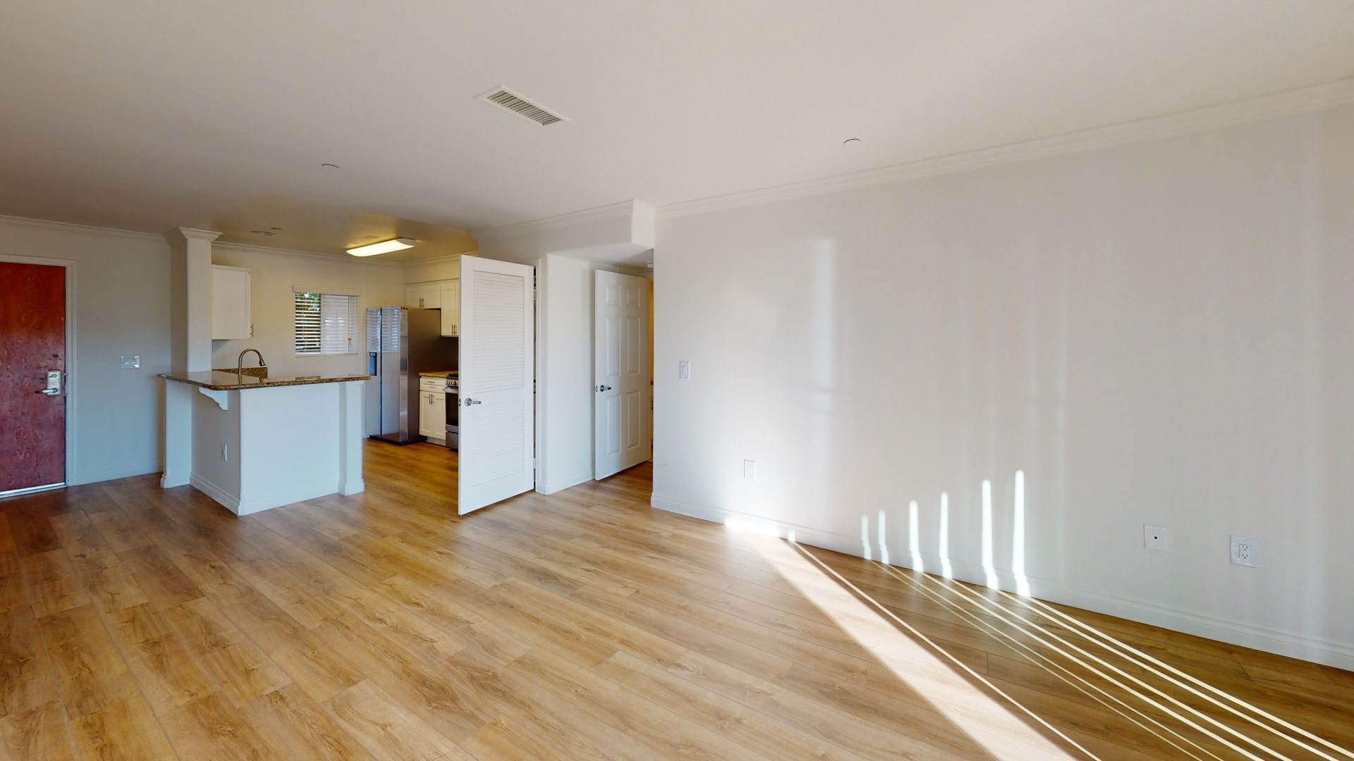 Bright, empty living space with wood flooring, white walls, and a partially visible kitchen.