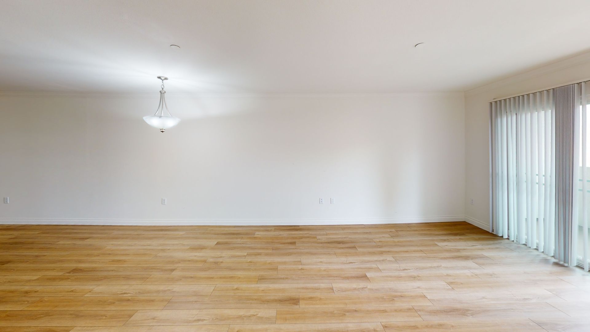 Empty room with light wood floor, white walls, and a glass door with curtains. A chandelier hangs from the ceiling.