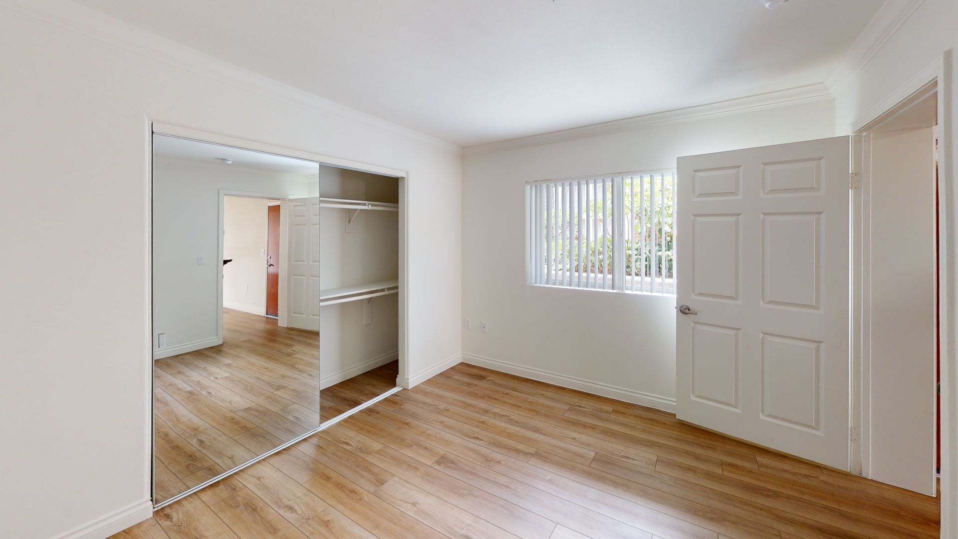 Empty bedroom with mirrored closet doors, wooden floors, and a closed white door.