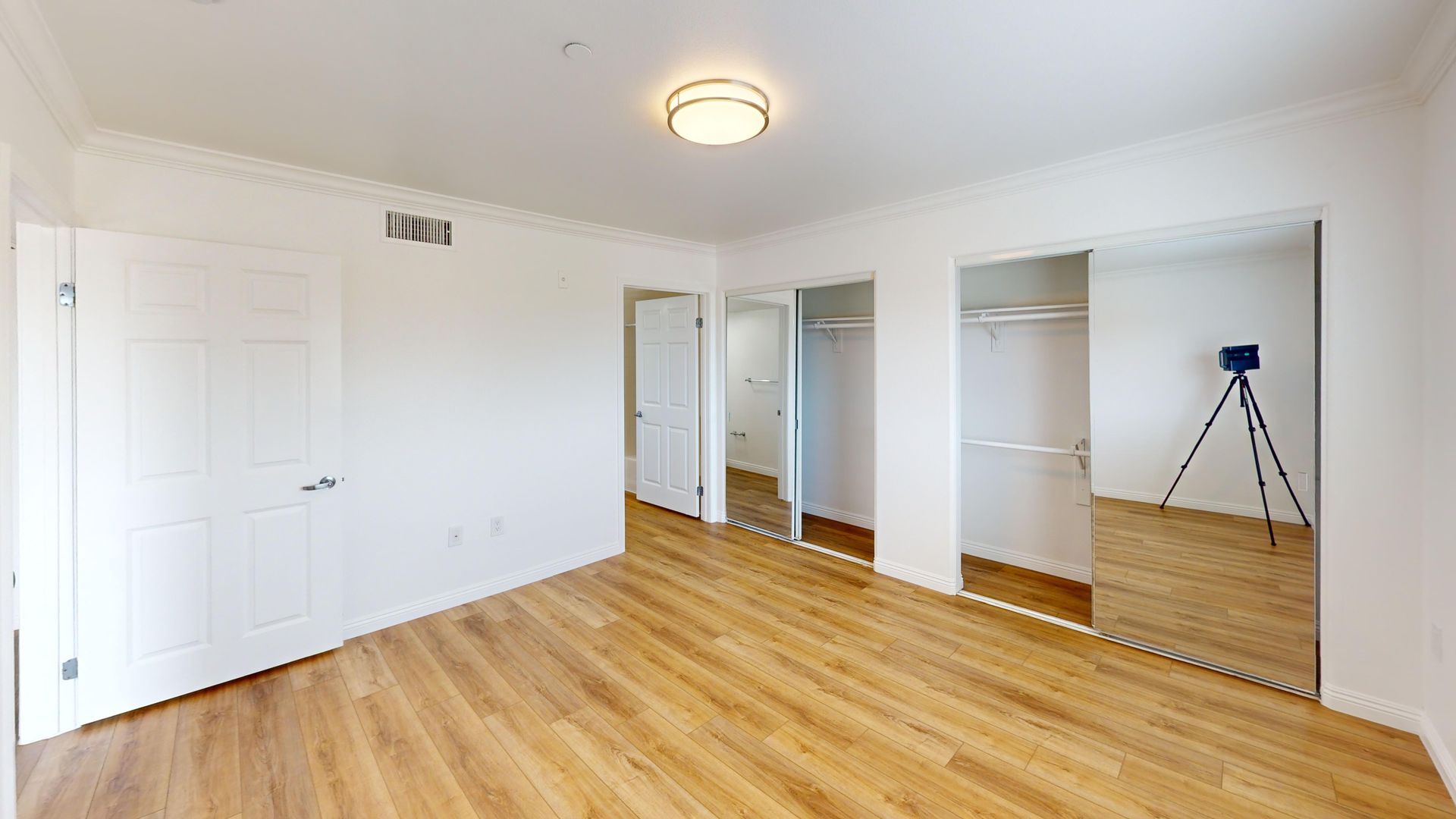 Empty room with hardwood floors, white walls, and mirrored closet doors. A tripod stands in front of the mirrors.