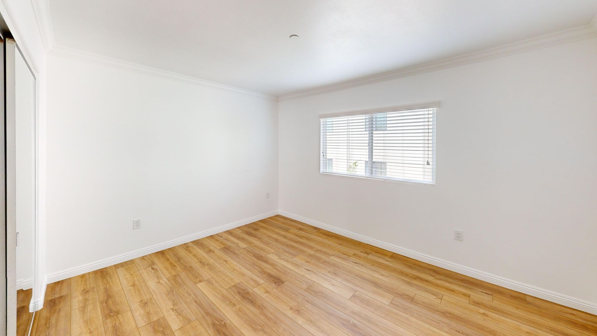 Empty bedroom with light wood flooring, white walls, and a window with blinds.