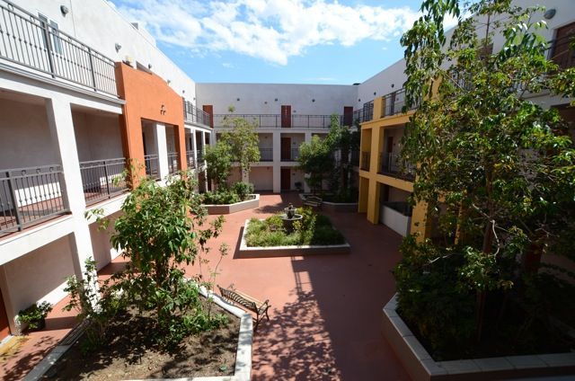 Courtyard with red tile floor, white and colorful buildings, greenery, and balconies under a blue sky.
