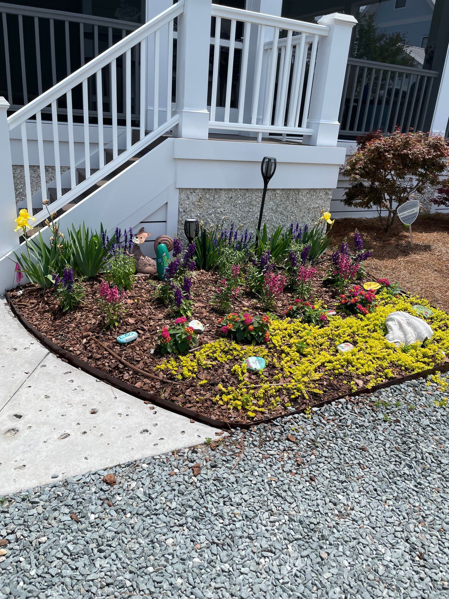Flowerbed with colorful flowers, wood chips, and stepping stone accents in front of a white porch.