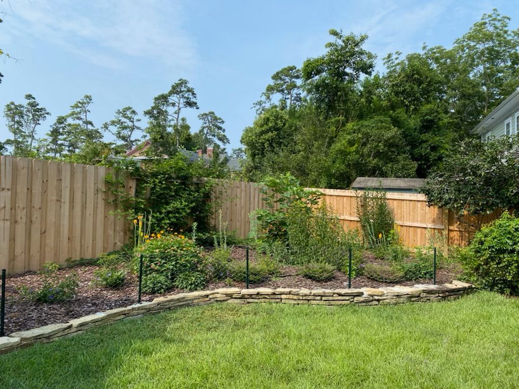 Lawn with a stone-edged garden bed against a wooden fence, with trees and a blue sky in the background.