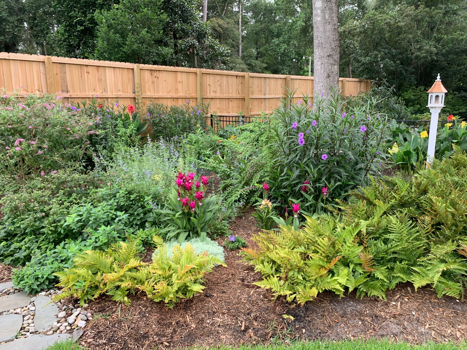 Lush garden bed with various plants, flowers, and a birdhouse against a wooden fence.