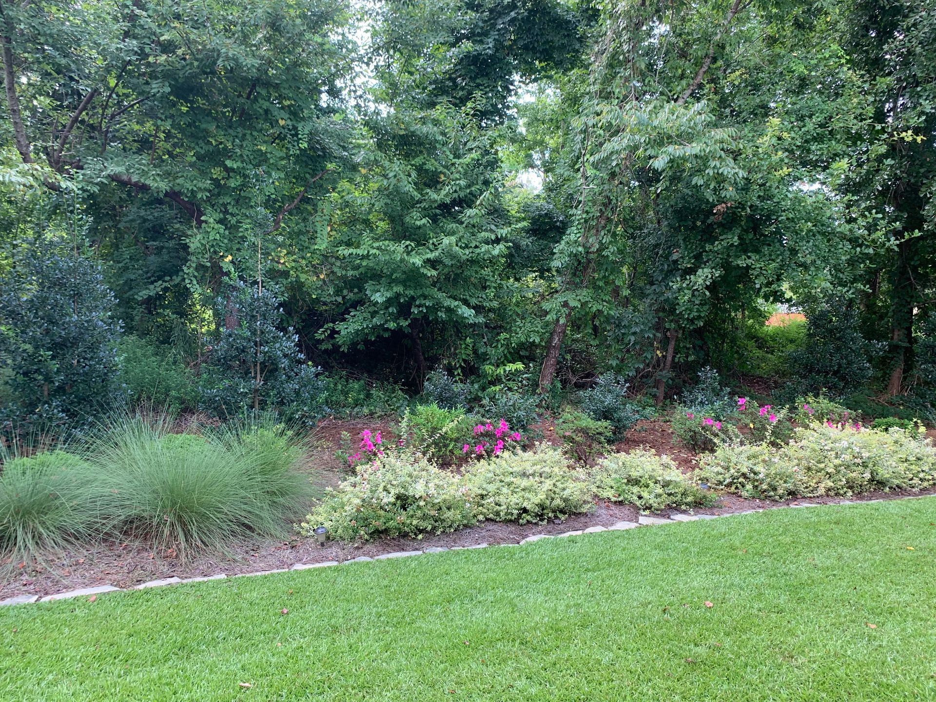 Green lawn borders a garden bed with bushes, flowering plants, and trees.