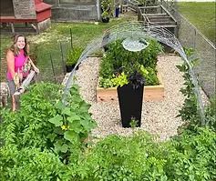 Woman in a garden with a raised bed, black planter, and metal arch, surrounded by green plants.