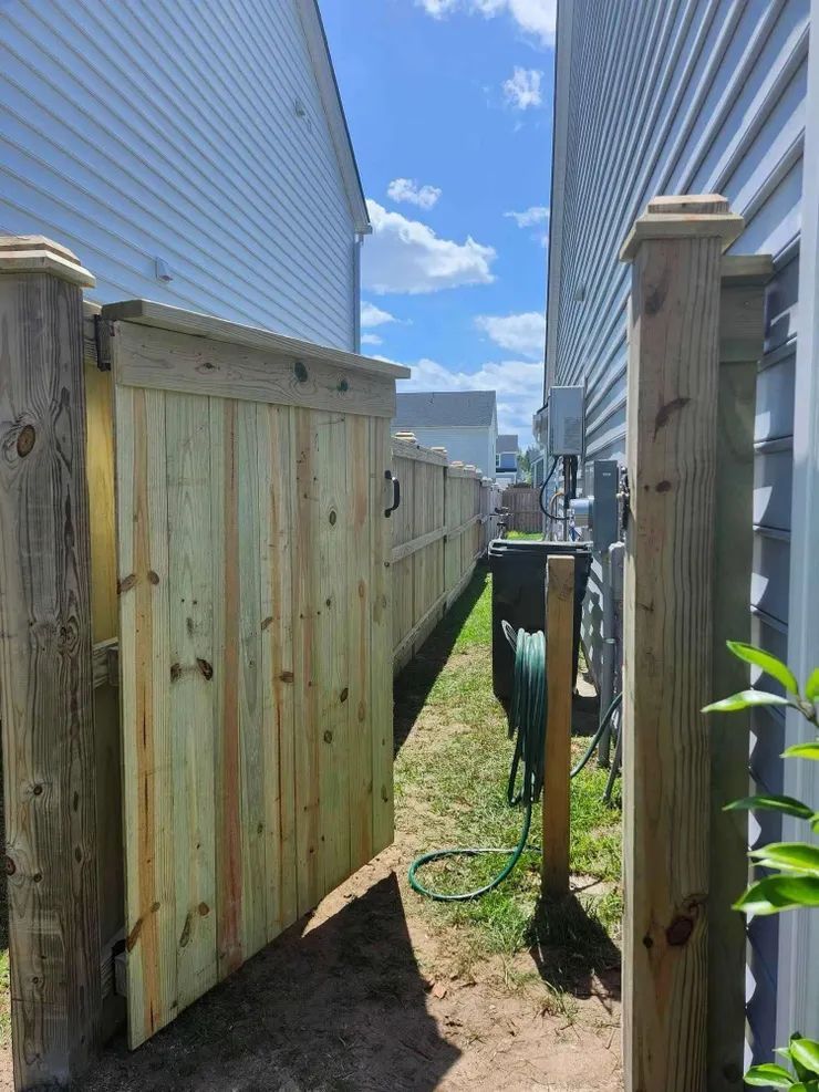 Wooden gate in a narrow passageway between two houses, with a long wooden fence extending into the distance on a sunny day.
