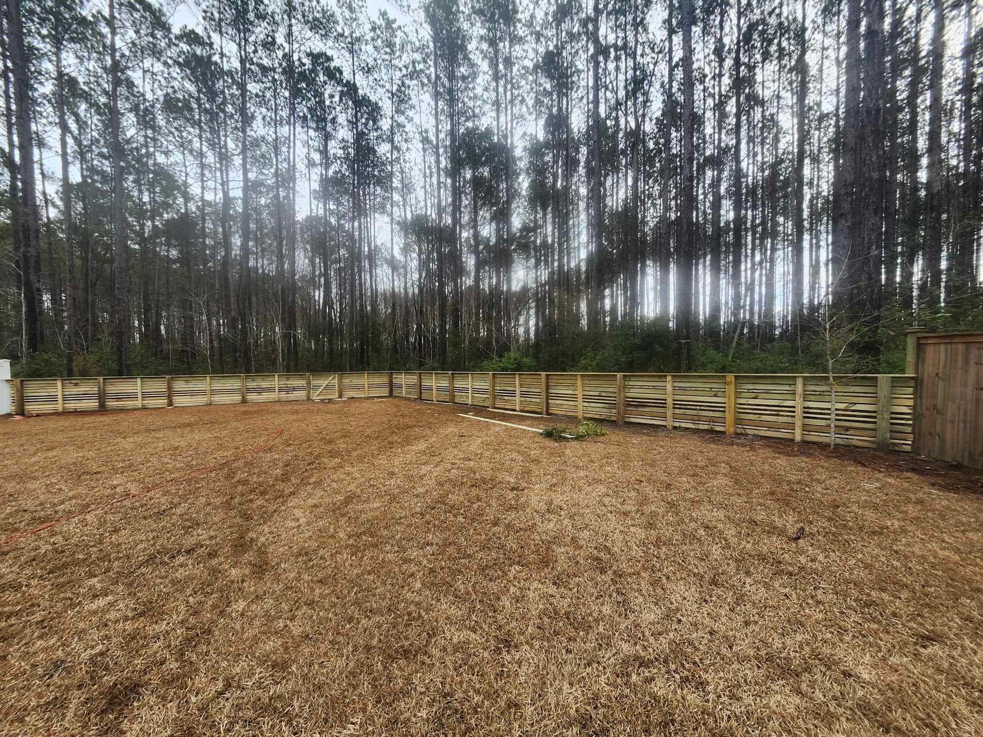 Wooden fence surrounding a gravel yard, with tall trees in the background under an overcast sky.