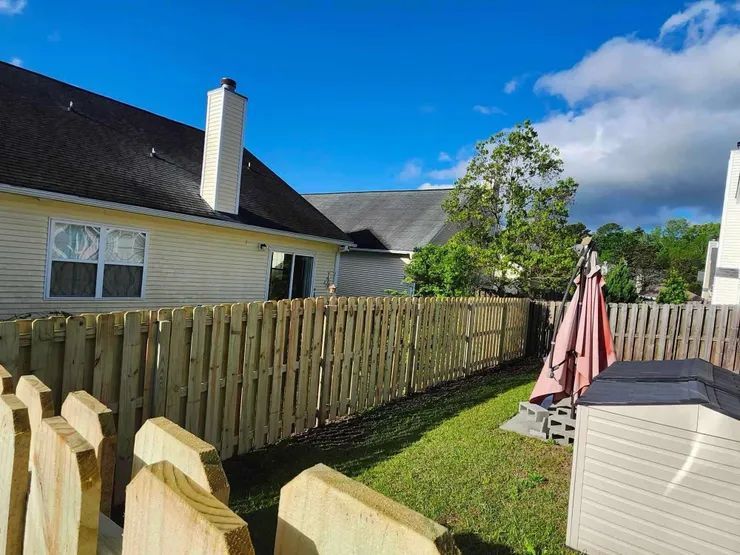 A newly constructed wooden fence surrounds a grassy backyard, with houses and a clear sky in the background.