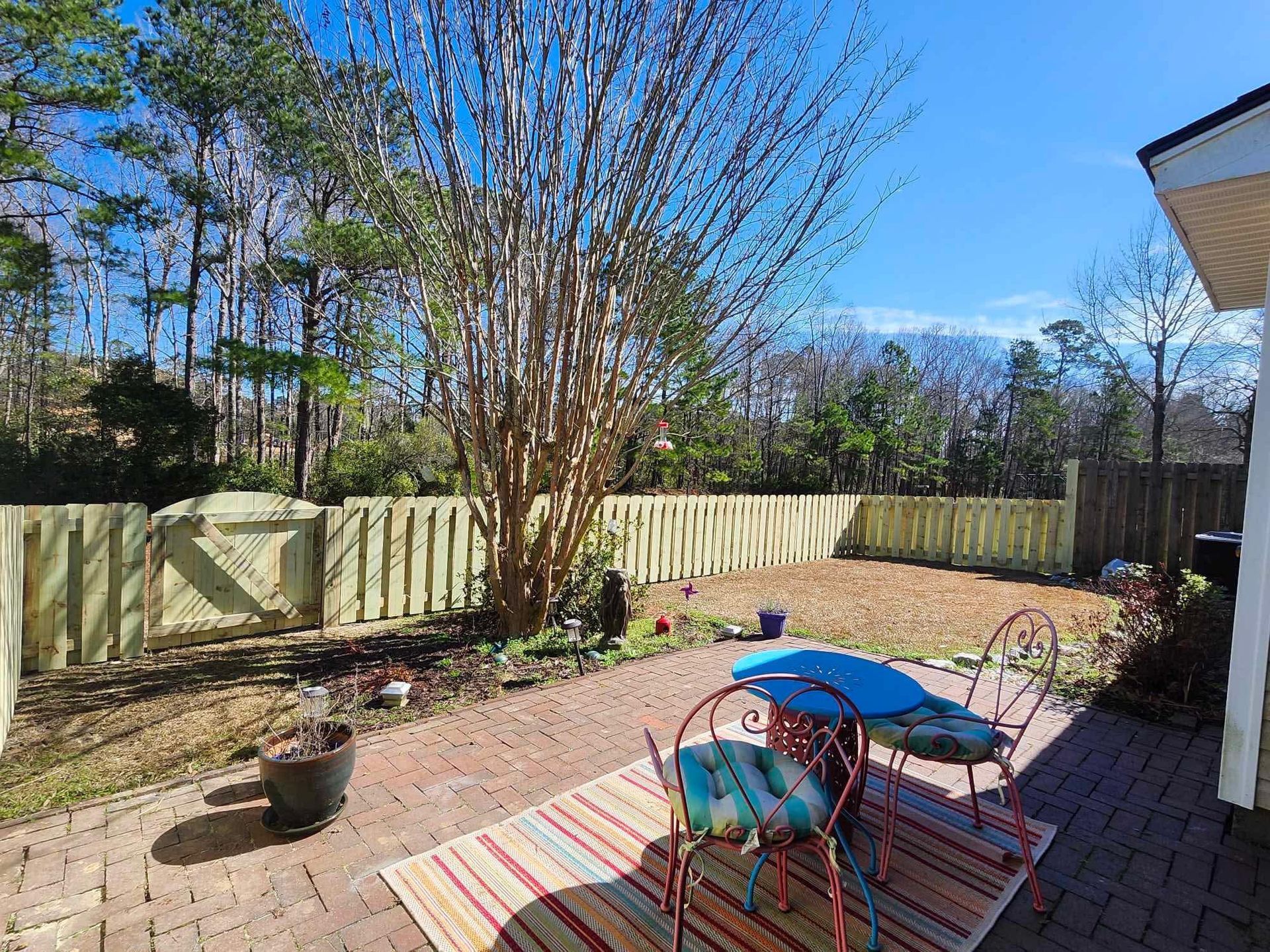 Backyard with brick patio, table and chairs, fenced yard, and bare tree under blue sky.