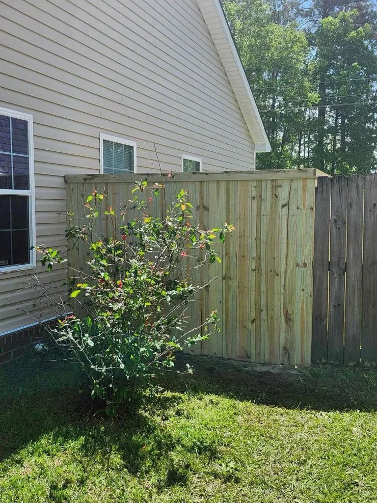 Wooden fence alongside a house, partially obscuring a bush in a grassy yard.