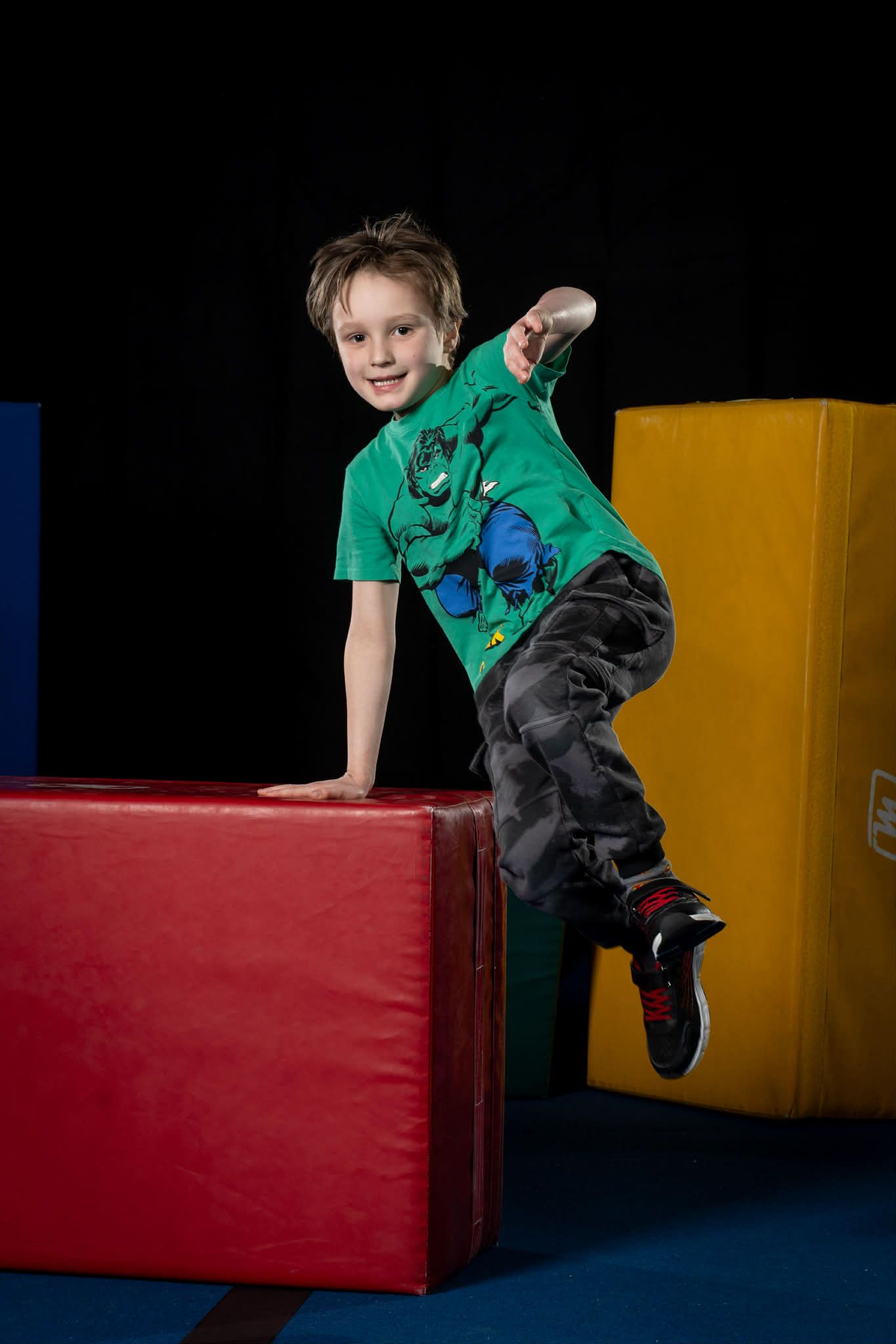 Un garçon en chemise verte saute par-dessus un bloc rembourré rouge dans un gymnase intérieur.