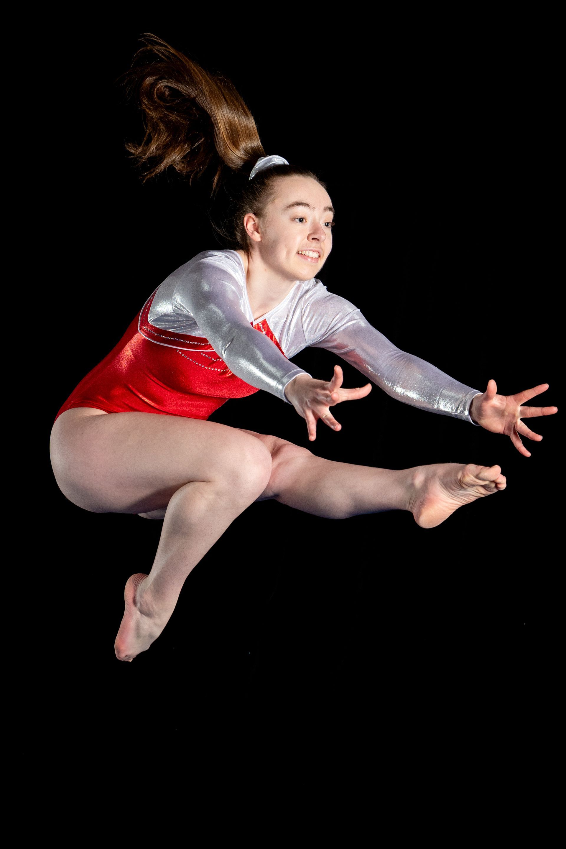 Une gymnaste en justaucorps rouge et argent saute sur fond noir, souriante, les cheveux attachés en queue de cheval.