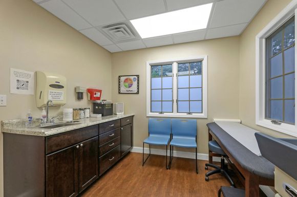 Medical examination room with exam table, sink, cabinets, and blue chairs.