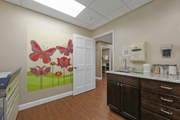 Medical exam room with butterfly art, sink, and open door to another room.