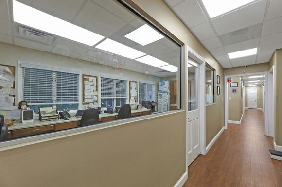 Hallway with wood floors and tan walls. Large window reveals office space. Fluorescent lights.