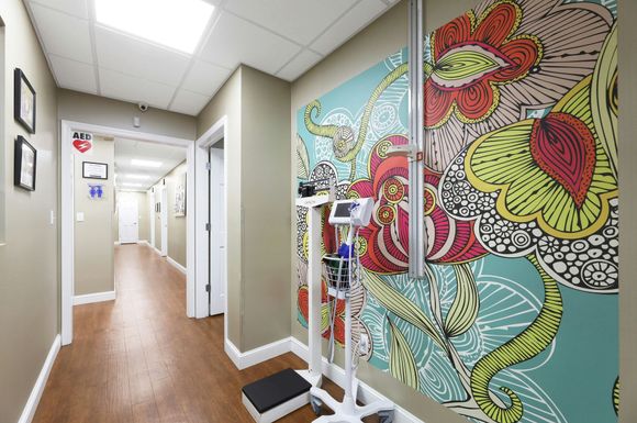 Hallway in a medical office with colorful floral mural, weight scale, and several closed doors.