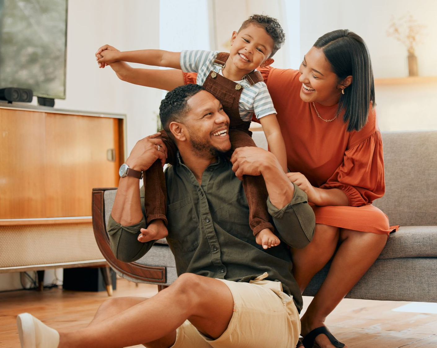 A man with a young boy on his shoulders  is sitting on the floor. A woman is sitting behind them and they are all smiling.