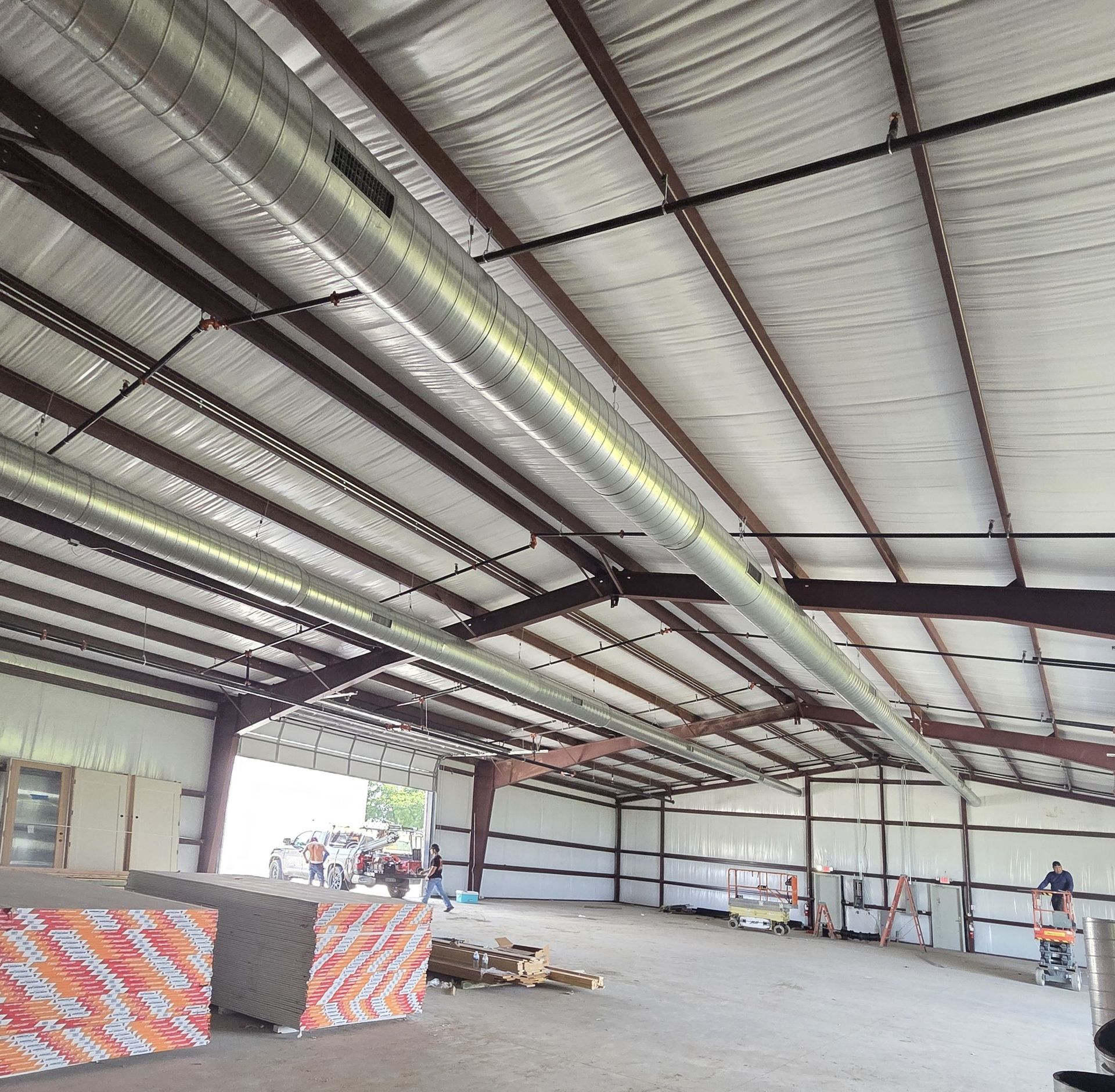 Interior of a large metal warehouse. Shiny ductwork runs across the ceiling. Building materials and workers are visible.