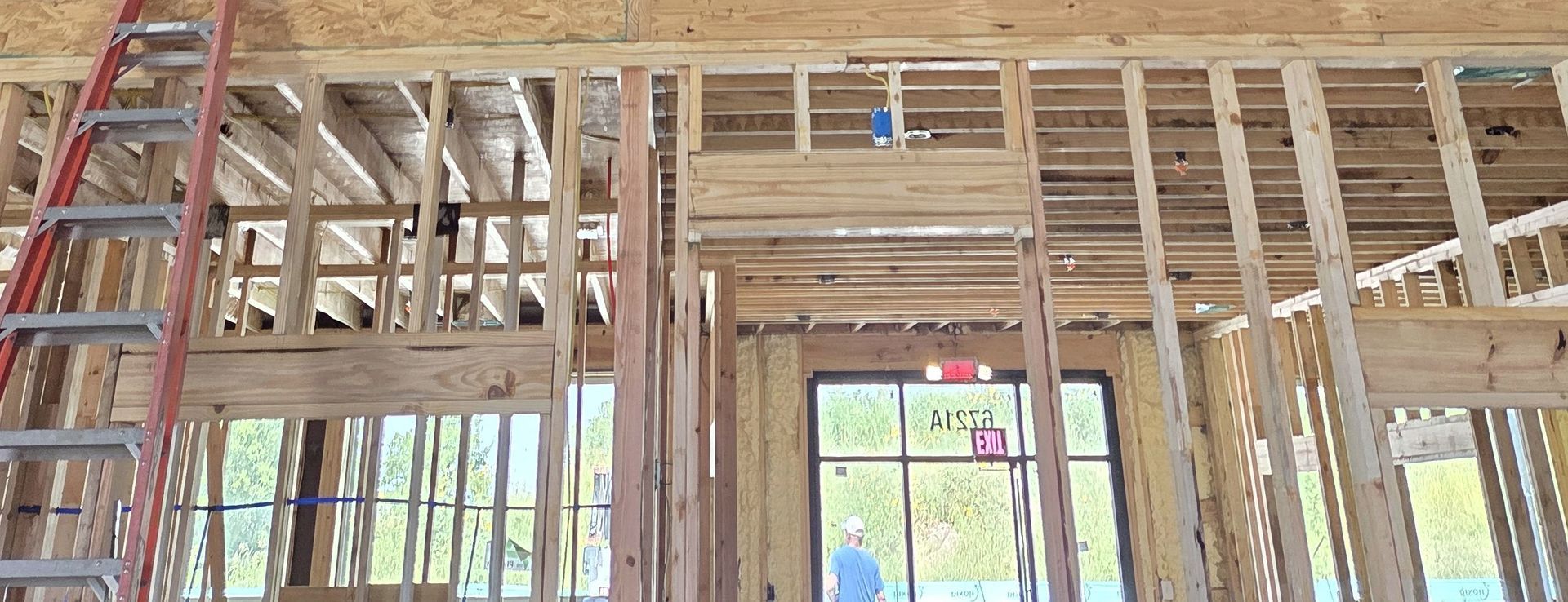 Interior view of a building under construction showing exposed wooden framing, a red ladder, and a doorway with a person visible through the glass.